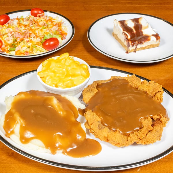 Chicken fried steak with gravy, mashed potatoes, mac and cheese, salad, and cheesecake on a wooden table.