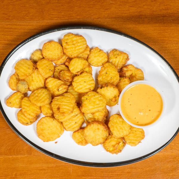 A white oval plate with a black rim holds fried pickle chips and a side of orange dipping sauce.