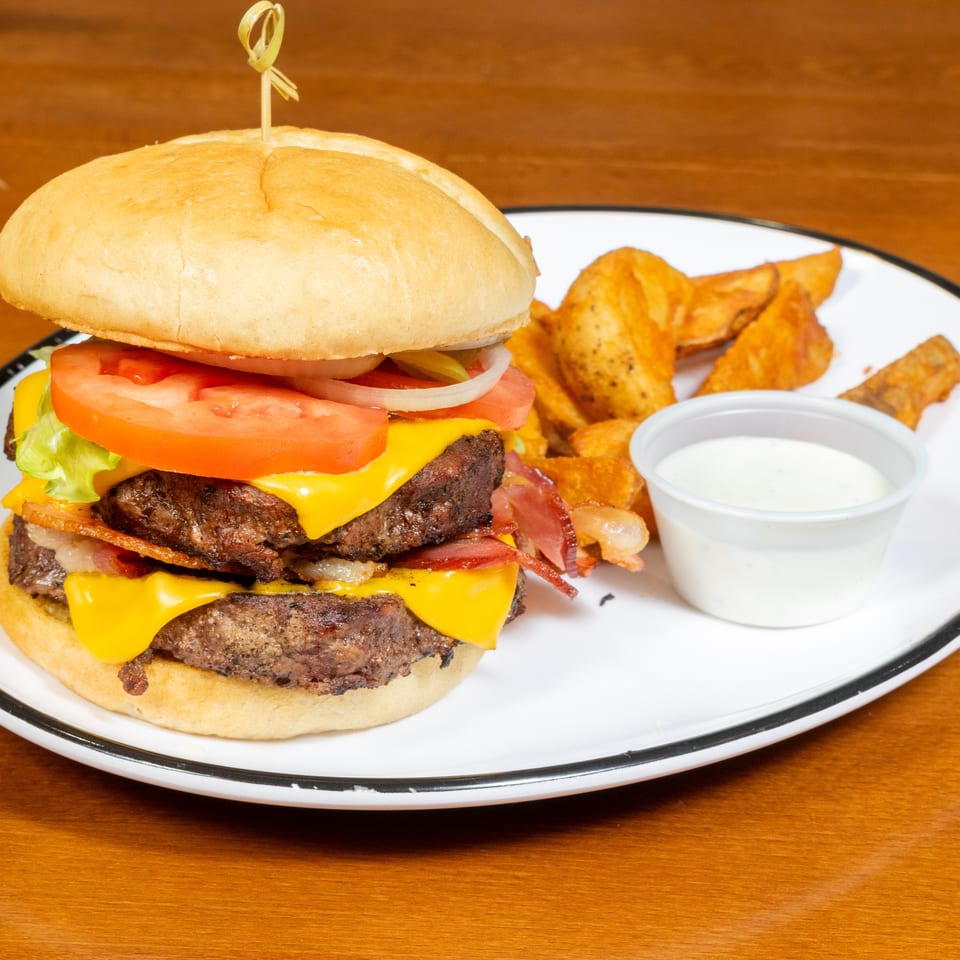Double cheeseburger with bacon, lettuce, tomato, onion, and a side of potato wedges and dip.
