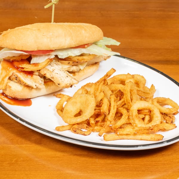 Grilled chicken sandwich with onion rings, lettuce, tomato, and BBQ sauce on a white plate.