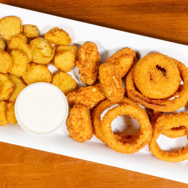 A white rectangular plate filled with golden-brown fried pickles, onion rings, and cheese curds, with a side of ranch.