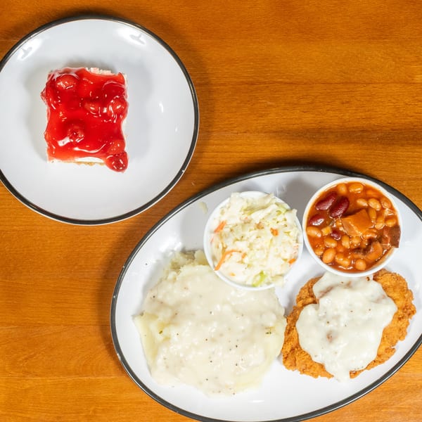 A plate of chicken fried steak with gravy, mashed potatoes, coleslaw, baked beans, and cherry dessert.