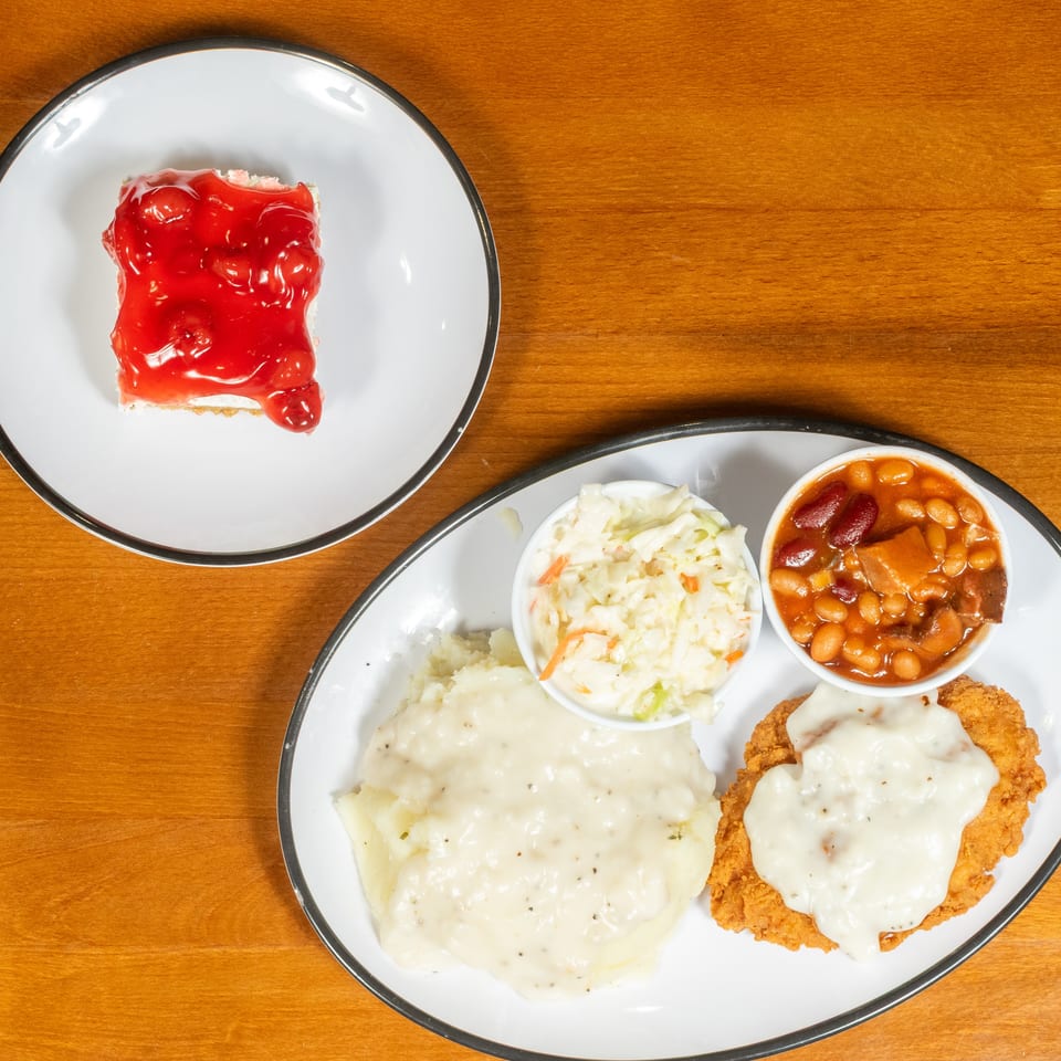 A plate of chicken fried steak with gravy, mashed potatoes, coleslaw, baked beans, and cherry dessert.