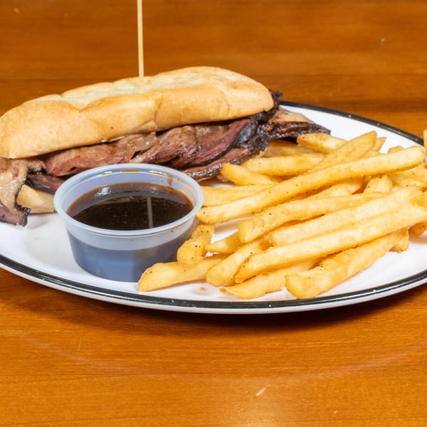 Brisket sandwich on a white plate with a side of fries and a small cup of au jus.