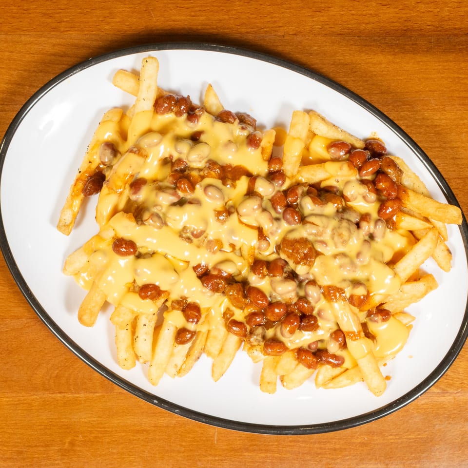 Chili cheese fries on a white oval plate, topped with melted yellow cheese and brown beans.